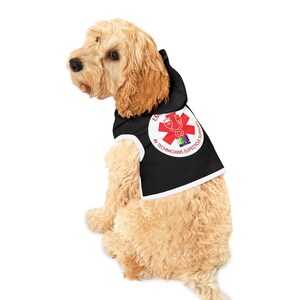 Puede incluir: Un perro marrón claro con una sudadera con capucha negra con ribete blanco. La sudadera tiene un emblema rojo y blanco con el texto "EM AV TECHNICIANS (LIFECYCLE SUPPORT)". El perro está sentado y mirando hacia la izquierda.