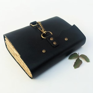 May include: A black leather journal with a brass clasp and rivets. The journal has a flap closure and a vintage look with aged paper pages. A small green leaf rests beside the journal.