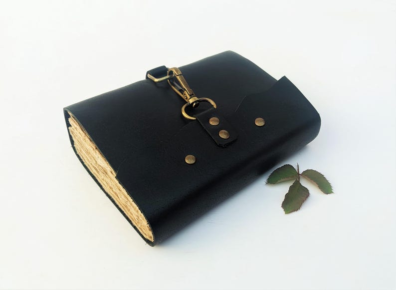 May include: A black leather journal with a brass clasp and rivets. The journal has a flap closure and a vintage look with aged paper pages. A small green leaf rests beside the journal.