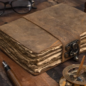 May include: A brown leather journal with a strap closure. The journal has aged, textured pages and a metal clasp. A pen, glasses, and a sundial are also visible, suggesting a vintage or antique theme.