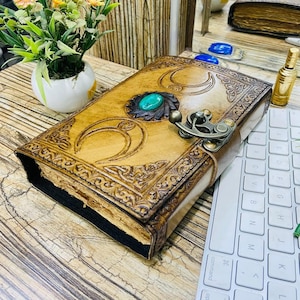 May include: A handcrafted leather journal with intricate embossed designs and a turquoise stone accent. The journal features a metal clasp closure and is displayed on a wooden surface. The cover has crescent moon and floral patterns.