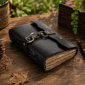May include: A black leather journal with a clasp closure and aged paper pages. The journal is adorned with brass hardware and embossed detailing. Two pens are next to the journal. The journal is on a wooden surface.