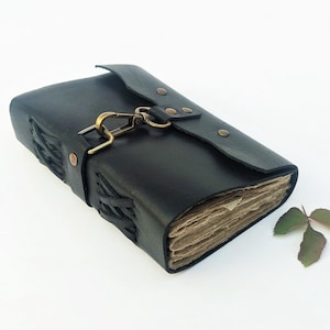 May include: Black leather journal with a clasp closure and stitched detailing. The journal has aged-looking pages and a decorative clasp. A small sprig of leaves rests beside the journal.