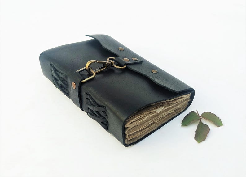 May include: Black leather journal with a clasp closure and stitched detailing. The journal has aged-looking pages and a decorative clasp. A small sprig of leaves rests beside the journal.