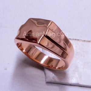 May include: A rose gold ring with a geometric design. The ring has a rectangular face with a slightly curved top and a smooth, polished finish. The text "ArtisanMagicCo" is visible on the ring.