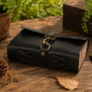 May include: A black leather journal with a flap closure and a brass clasp. The journal has stitched detailing on the front and aged paper pages. A pen and a pinecone are on the wooden surface.