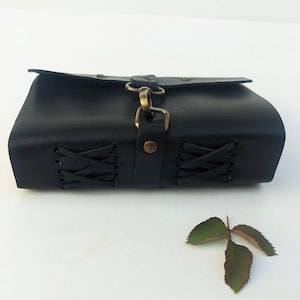 May include: A black leather pouch with a flap closure and brass-colored hardware. The sides of the pouch feature decorative lacing. A small, three-leaf sprig rests near the pouch.