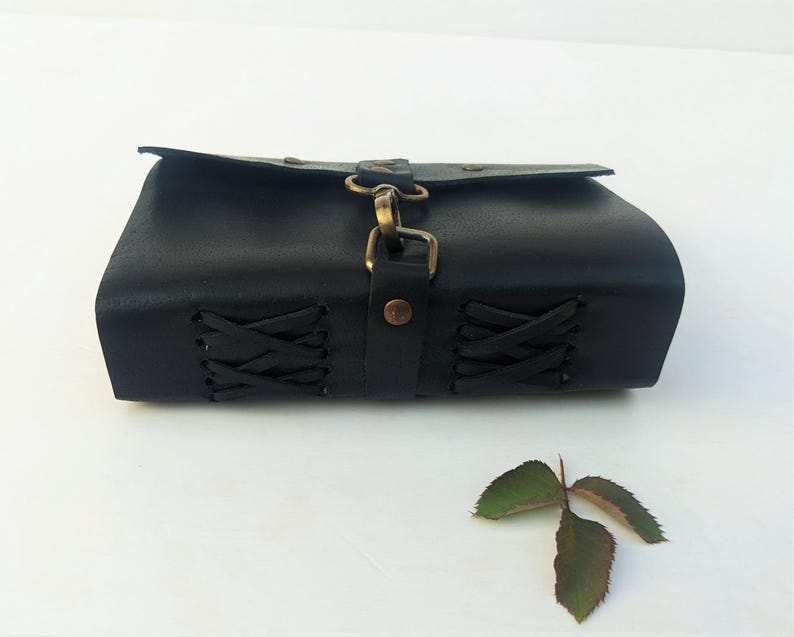 May include: A black leather pouch with a flap closure and brass-colored hardware. The sides of the pouch feature decorative lacing. A small, three-leaf sprig rests near the pouch.