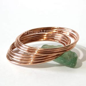 May include: A stack of copper-colored wire rings, possibly for jewelry making, resting on a green stone against a white background. The rings are tightly coiled and appear to be made of a thin, flexible metal. The text "ArtisanMagicCo" is visible.