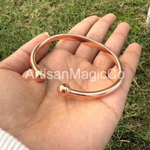 May include: A copper-colored cuff bracelet with rounded ball ends. The bracelet is held in a hand against a background of green grass. The text "ArtisanMagicCo" is visible on the bracelet.