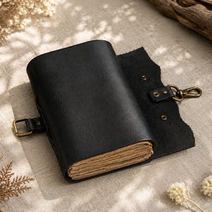May include: A black leather journal with a brass buckle closure and a matching clasp. The journal is open, revealing aged, cream-colored pages. The cover is textured, and the journal is lying on a neutral-toned surface.