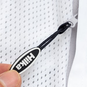 Puede incluir: Un tirador de cremallera blanco con un cordón negro y una etiqueta ovalada negra que dice "HIIKA" en letras blancas.