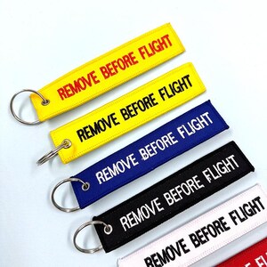 Könnte beinhalten: Eine Sammlung rechteckiger Schlüsselanhänger in Gelb, Blau, Schwarz und Weiß, jeweils mit einem silbernen Schlüsselring. Die Schlüsselanhänger zeigen den Text "REMOVE BEFORE FLIGHT" in kontrastierenden Farben. Die Schlüsselanhänger sind für Luftfahrt-Enthusiasten konzipiert.
