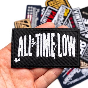 Peut inclure: Un écusson rectangulaire noir avec l'inscription blanche brodée "ALL TIME LOW" et un petit graphisme. L'écusson est tenu dans une main, avec d'autres écussons visibles en arrière-plan.