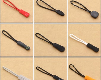 Könnte beinhalten: Neun Reißverschluss-Zipper mit verschiedenen Farben für Kordel und Endkappen. Die Kordeln sind schwarz, weiß, rot und orange. Die Endkappen sind schwarz, grau und orange. Einige Endkappen haben Text, darunter "PRODP" und "RA".