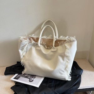 Può includere: Borsa tote in tela color crema con frange sulla parte superiore e sui lati. Ha due manici e un interno spazioso. La borsa è appoggiata su una giacca di pelle nera e una rivista.