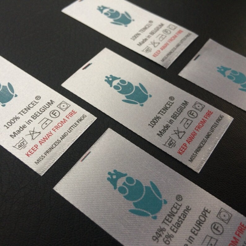 May include: Close-up of four white fabric labels with a frog logo and text that reads "100% TENCEL&reg; Made in BELGIUM 40&deg; KEEP AWAY FROM FIRE MISS PRINCESS AND LITTLE FROG" and "94% TENCEL&reg; 6% Elastane in EUROPE KEEP AWAY FROM FIRE MISS PRINCESS AND LITTLE FROG".