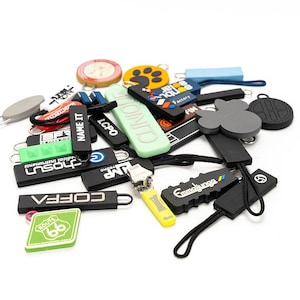 May include: A collection of various zipper pulls in different shapes, sizes, and colors. Some pulls have text like "NAME IT", "CLINIQUE", and "ROUTE 66". The pulls are made of rubber and plastic, with black cords attached. The background is white.