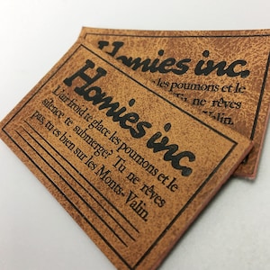 Könnte beinhalten: Zwei braune Leder-Patches mit schwarzem Text. Der obere Patch sagt "Homies inc." und der untere Patch sagt "Homies inc. L'air froid te glace les poumons et le silence te submerge? Tu ne rêves pas, tu es bien sur les Monts-Valin."