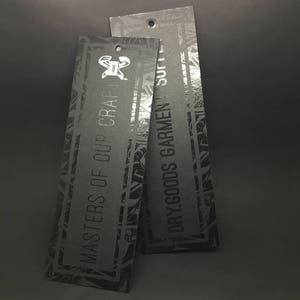 Pode incluir: Duas etiquetas pretas com uma borda de padr&atilde;o geom&eacute;trico. A etiqueta superior diz "DRY GOODS GARMENT" e a etiqueta inferior diz "MASTERS OF OUR CRAFT".