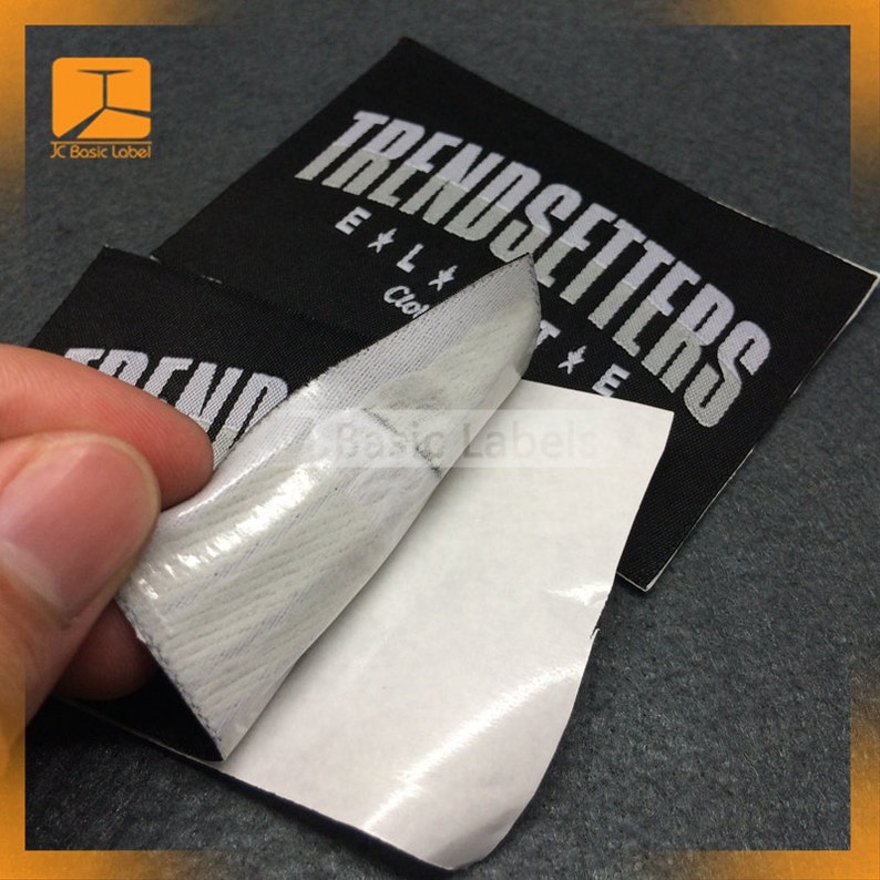 May include: Black fabric label with white text that reads "TRENDSETTERS" with a white star on either side of the word "SETTERS". The label has a white adhesive backing.