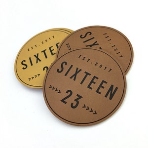 Könnte beinhalten: Drei runde Leder-Patches mit dem Text "SIXTEEN EST. 2017" und der Zahl "23" in schwarzer Schrift. Die Patches sind braun und gold.
