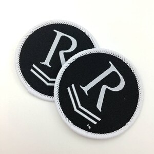 May include: Two black and white embroidered patches with a white letter "R" and a white chevron design. The patches have a white stitched border.