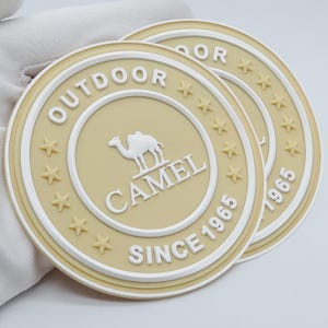Puede incluir: Dos posavasos redondos beige con texto blanco y un logotipo de camello. El texto dice "OUTDOOR CAMEL SINCE 1965" y está dispuesto alrededor del borde de los posavasos. Los posavasos tienen un patrón de estrellas.