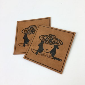 Könnte beinhalten: Zwei braune Lederpatches mit schwarzen Tintenillustrationen einer Person, die einen Hut trägt und ein Messer hält. Der Text "Papi Gang" ist unten auf dem Patch aufgedruckt.