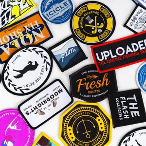 Op de afbeelding: Een assortiment van kleurrijke geborduurde patches met verschillende ontwerpen en tekst, waaronder "The Flash Collective", "Fresh Baits", "Take The Reins", "Uploaded The Online Fishing Series", "Fly Shop NOLA", "Moodricht's", "Palm", "Icicle Brewing Company" en "Naomh Mearnós".