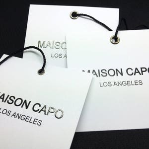 Pode incluir: Tr&ecirc;s etiquetas brancas com texto preto e letras em folha prateada. As etiquetas dizem "Maison Capo Los Angeles". Cada etiqueta tem um cord&atilde;o preto amarrado com um ilh&oacute; de metal prateado.