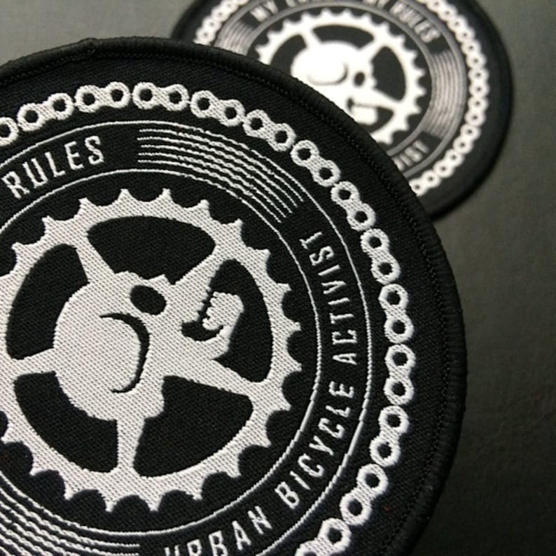 May include: Black and white embroidered patch with a gear design and the text "Urban Bicycle Activist" and "Rules" around the edge.