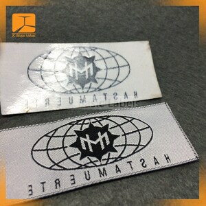 May include: Two white fabric labels with black stitching around the edges. The labels feature a black and white logo of a stylized "MM" inside a circle with a globe design. The text "ETREUMAT2AH" is printed on the labels in a mirrored font.