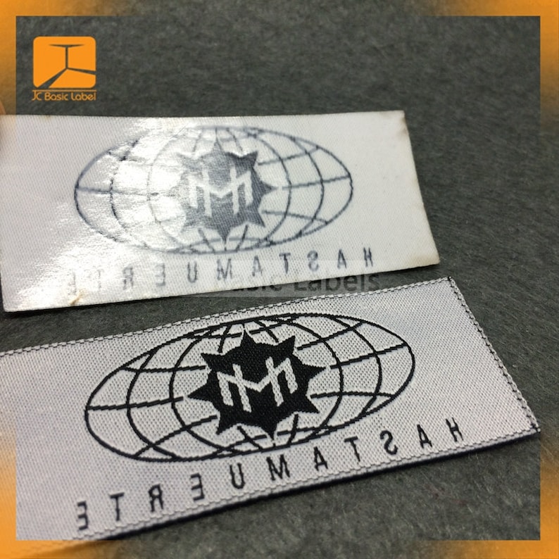 May include: Two white fabric labels with black stitching around the edges. The labels feature a black and white logo of a stylized "MM" inside a circle with a globe design. The text "ETREUMAT2AH" is printed on the labels in a mirrored font.