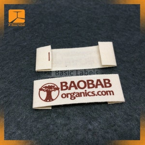Pode incluir: Duas etiquetas de tecido branco sujo. Uma etiqueta est&aacute; dobrada com uma pequena costura vermelha. A outra etiqueta est&aacute; desdobrada. As etiquetas t&ecirc;m o texto "BAOBAB organics.com" em castanho e um gr&aacute;fico de &aacute;rvore. O fundo &eacute; tecido cinzento escuro.
