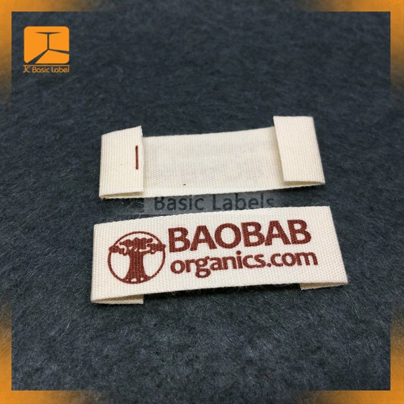 Pode incluir: Duas etiquetas de tecido branco sujo. Uma etiqueta est&aacute; dobrada com uma pequena costura vermelha. A outra etiqueta est&aacute; desdobrada. As etiquetas t&ecirc;m o texto "BAOBAB organics.com" em castanho e um gr&aacute;fico de &aacute;rvore. O fundo &eacute; tecido cinzento escuro.