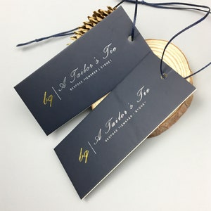 Könnte beinhalten: Zwei rechteckige Anhänger in Dunkelblau mit abgerundeten Ecken. Die Anhänger haben einen weißen Rand und goldene Beschriftung mit dem Text "A Tailor's Tie" und "BESPOKE TIEMAKER | SYDNEY".