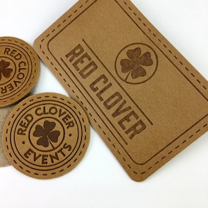 Könnte beinhalten: Hellbraune Lederaufnäher und Untersetzer mit dem Text "RED CLOVER" und einem vierblättrigen Kleeblatt-Design. Der rechteckige Aufnäher hat einen genähten Rand. Zwei runde Untersetzer zeigen ebenfalls den Text und das Kleeblatt-Design.