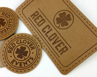 May include: Tan leather patches and coasters with the text "RED CLOVER" and a four-leaf clover design. The rectangular patch has a stitched border. Two round coasters also feature the text and clover design. The items are likely for branding or crafts.