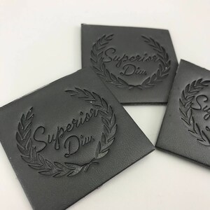 Könnte beinhalten: Drei schwarze Leder-Etiketten mit einem Lorbeerkranz-Design und dem Text "Supervisor Div." in der Mitte geprägt.
