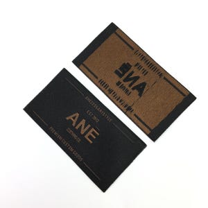 May include: Two rectangular clothing labels. One is black with the text "ANE CLOTHING CO." and "PREMIUM CUSTOM GOODS." The other label is brown with the text "ANE." Both labels have a black border and are on a white background.