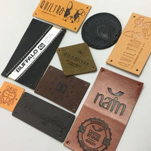 Könnte beinhalten: Sortiment aus Lederpatches mit verschiedenen Logos und Texten, darunter "Quiltro", "Buffalo", "Wrangler", "Quiksilver", "naim" und "Chicks That Rip".