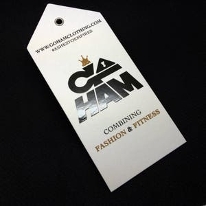 Pode incluir: Uma etiqueta branca com um logotipo prateado que diz "G.O.H.A.M" com uma coroa acima do "G". O texto "www.gohamclothing.com" e "#ashes to empires" est&aacute; impresso em preto no topo da etiqueta. O texto "Combining Fashion & Fitness" est&aacute; impresso em dourado na parte inferior da etiqueta.