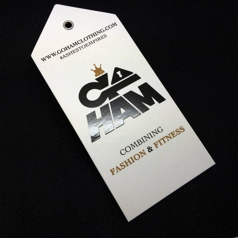 Pode incluir: Uma etiqueta branca com um logotipo prateado que diz "G.O.H.A.M" com uma coroa acima do "G". O texto "www.gohamclothing.com" e "#ashes to empires" est&aacute; impresso em preto no topo da etiqueta. O texto "Combining Fashion & Fitness" est&aacute; impresso em dourado na parte inferior da etiqueta.