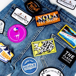 May include: A pair of blue jeans with various embroidered patches. The patches feature different designs, including a mountain range, a fly shop, a brewery, a gymnastics award, and a band logo. The patches are all sewn onto the jeans in a random pattern.