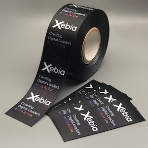 Può includere: Un rotolo di nastro nero con la parola "Xebia" e il testo "Creating Digital Leaders in the AI Era" stampato in bianco, blu, rosso e verde. Sono visibili anche diversi pezzi tagliati dello stesso nastro.