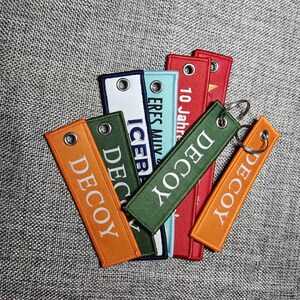 Könnte beinhalten: Eine Sammlung bestickter, rechteckiger Schlüsselanhänger in verschiedenen Farben, darunter Orange, Grün, Rot und Blau. Jeder Anhänger trägt das Wort "DECOY" oder einen anderen Text in weißer Schrift, mit einer Metallöse zur Befestigung am Schlüsselring.