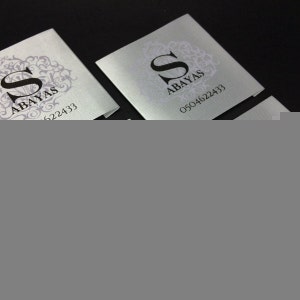 May include: Two silver business cards with a black letter "S" in a decorative frame and the text "Abayas" and a phone number.