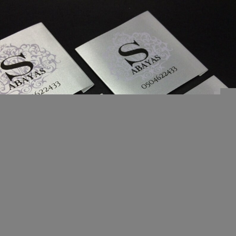 May include: Two silver business cards with a black letter "S" in a decorative frame and the text "Abayas" and a phone number.