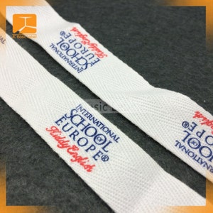 Pode incluir: Close-up de duas etiquetas de tecido branco com o texto "INTERNATIONAL SCHOOL OF EUROPE Kiddy English" impresso em azul e vermelho. As etiquetas t&ecirc;m uma textura tecida e est&atilde;o sobre uma superf&iacute;cie cinza escuro.